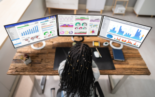 Photo of three computer screens displaying colourful data charts. A person with long dark hair is in the foreground with their back to the camera. They are looking at the screens. They are sitting at a desk.