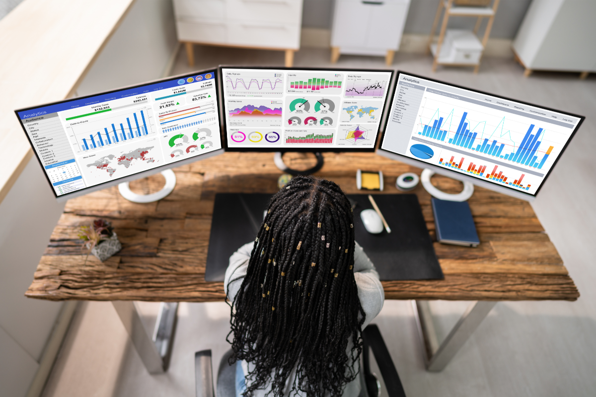 Photo of three computer screens displaying colourful data charts. A person with long dark hair is in the foreground with their back to the camera. They are looking at the screens. They are sitting at a desk.