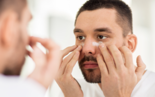 Young man applying cream under his eyes. He is looking in the mirror.