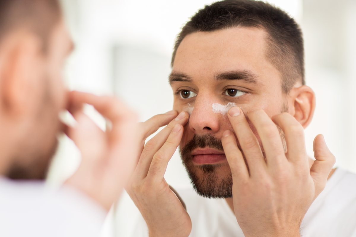 Young man applying cream under his eyes. He is looking in the mirror.