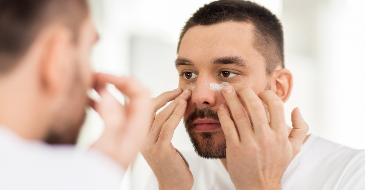 Young man applying cream under his eyes. He is looking in the mirror.